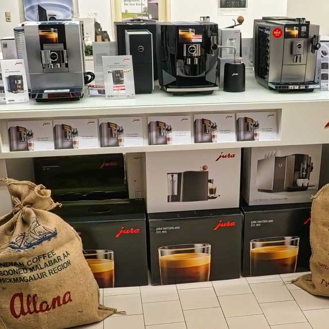 Regal mit drei modernen Jura-Kaffeemaschinen, jeweils mit Infokarte davor. Darunter gestapelte Produktkartons mit Jura-Logo und Abbildungen