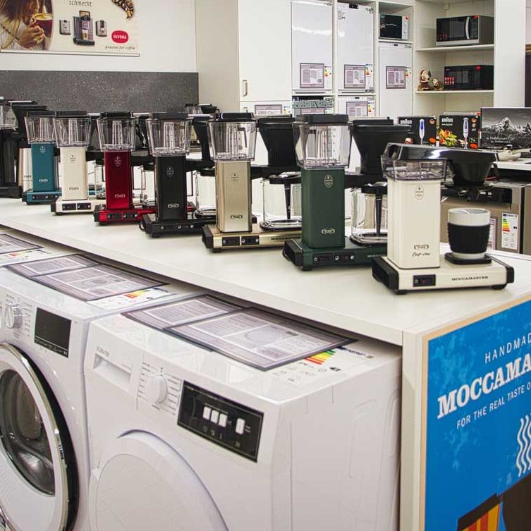 Verschiedene Moccamaster Kaffeemaschinen in verschiedenen Farben auf einem Verkaufstisch. Darunter Waschmaschinen. Im Hintergrund Mikrowelle